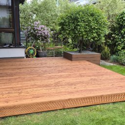 Drew-Tech Technikum Drewna - Zrealizowany taras z desek o ciepłym odcieniu drewna, otoczony zielonym trawnikiem i roślinnością. Widoczny fragment domu z oknem i elementami dekoracyjnymi.