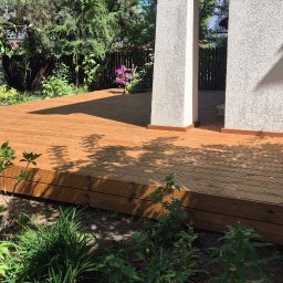 Drew-Tech Technikum Drewna - Nowoczesny taras z drewna, z subtelnym oświetleniem, wkomponowany w bujną zieleń ogrodu. Widoczne deski tarasowe i fragment budynku.