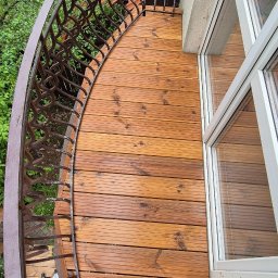 Drew-Tech Technikum Drewna - Kątowe ujęcie drewnianego tarasu balkonowego z metalową, dekoracyjną balustradą. Widoczne deski tarasowe i fragment okna. Na podłodze widoczne stopy osoby robiącej zdjęcie.