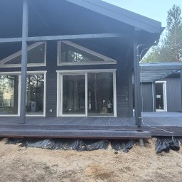 Drew-Tech Technikum Drewna - Nowoczesny dom z ciemnego drewna z rozległym tarasem. Widoczne duże okna i otoczenie leśne. Taras w trakcie wykończenia, z widoczną folią ochronną.