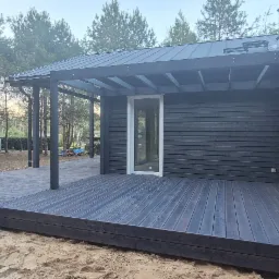 Ciemny, drewniany taras przylegający do nowoczesnego domu z ciemną elewacją i pergolą. W tle las sosnowy i piaszczyste podłoże.