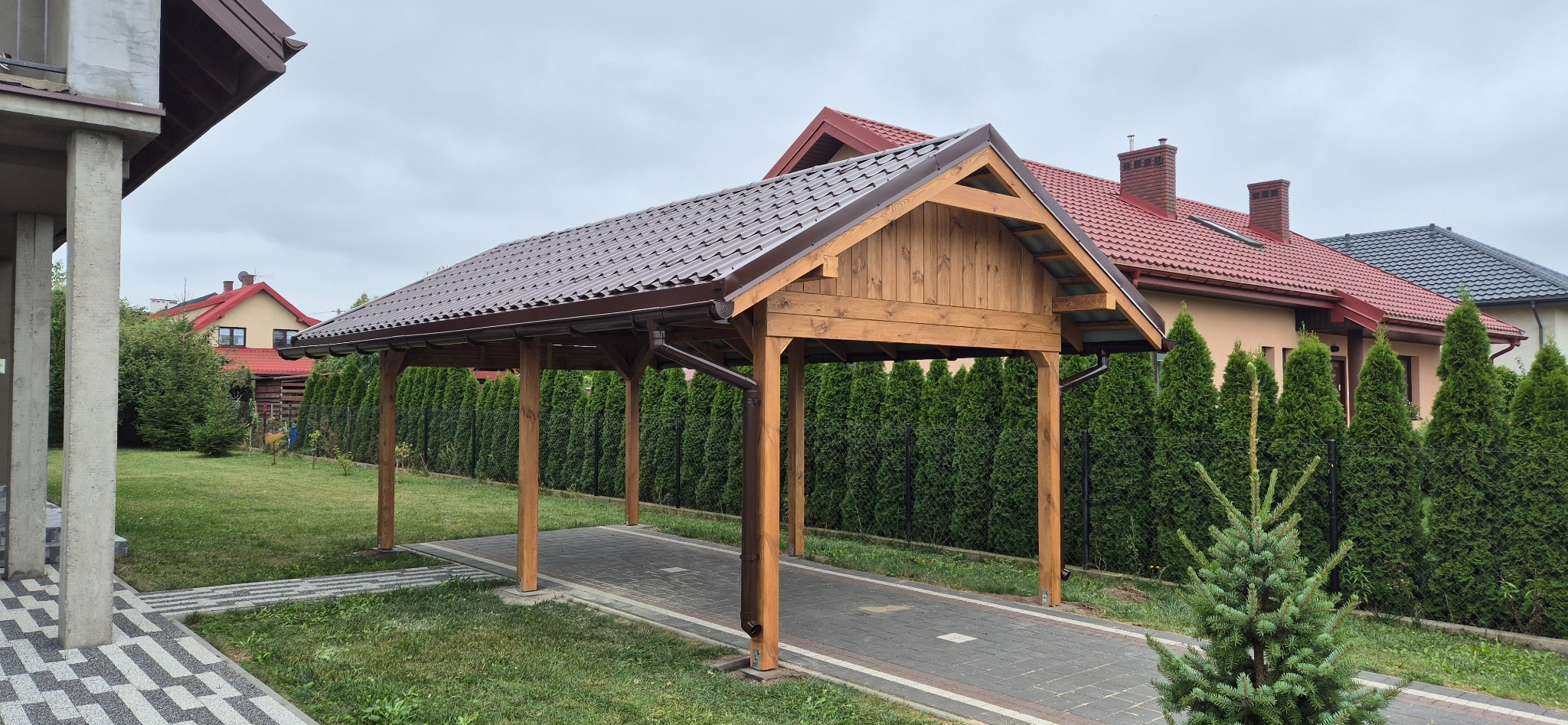 Drewniana wiata garażowa z brązowym dachem na posesji z tujami w tle, widok z boku.