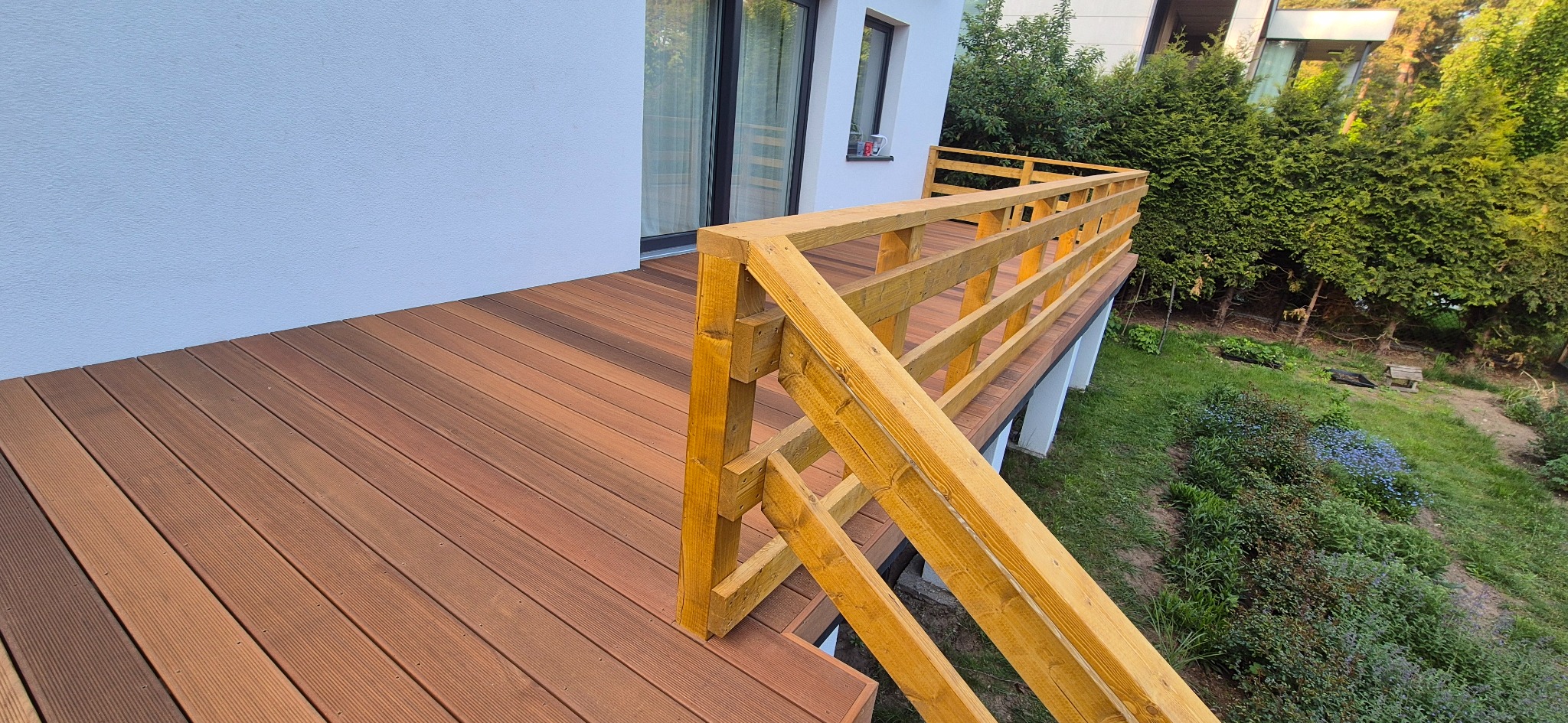 Kompozytowy taras z drewnianą balustradą, widok z boku, zieleń w tle. Deski w odcieniach brązu, pionowe elementy balustrady w kolorze jasnego drewna.
