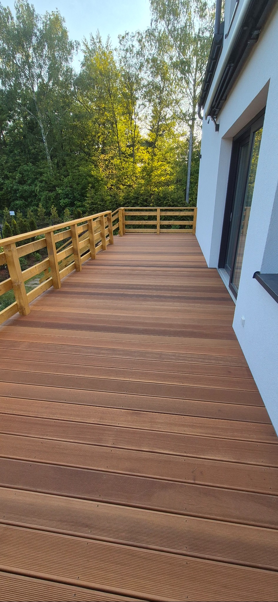 Nowoczesny taras z desek kompozytowych w odcieniach brązu, z drewnianą balustradą. Widok z góry na taras przylegający do białego budynku z oknem.