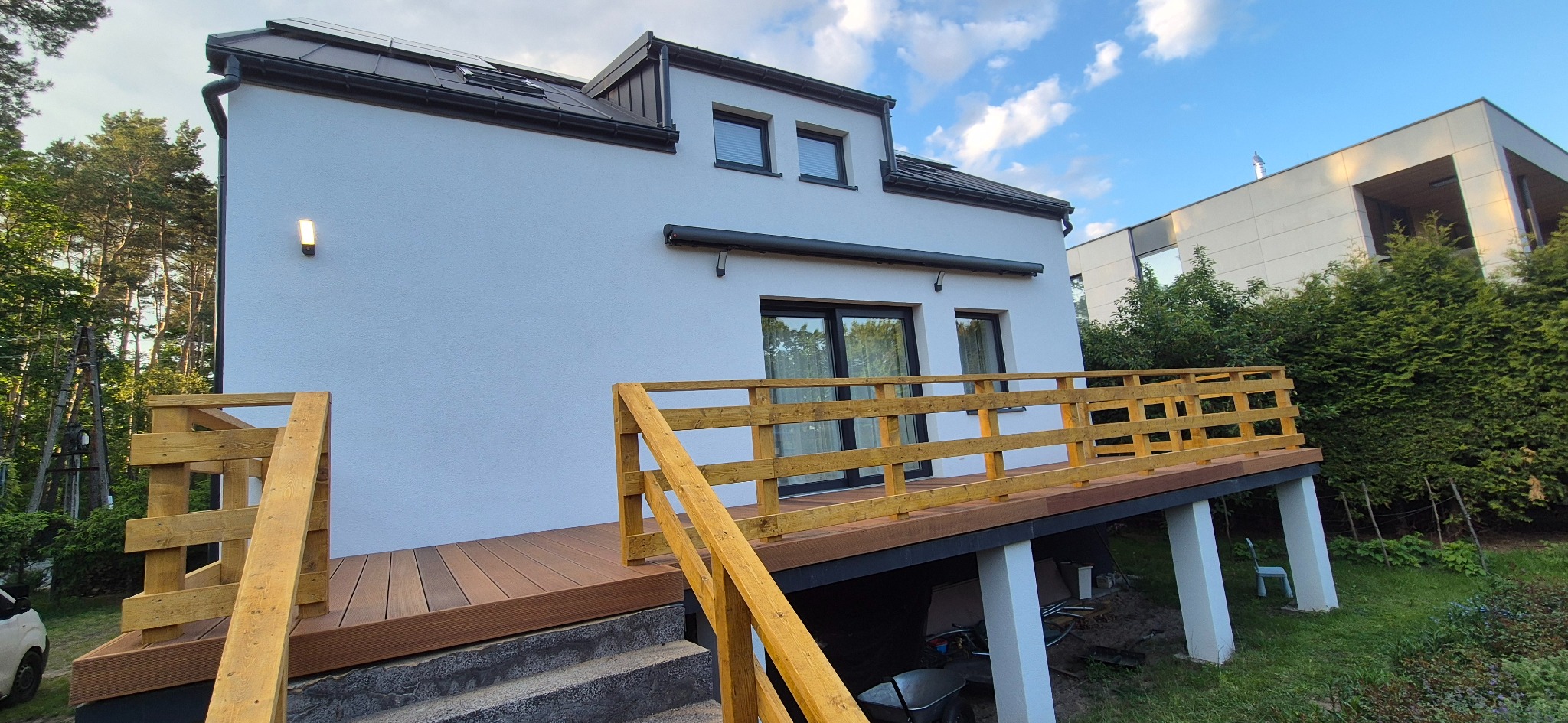 Nowoczesny taras z deski kompozytowej przy białym domu z drewnianą balustradą, widok z boku, z elementami ogrodu i błękitnym niebem w tle.