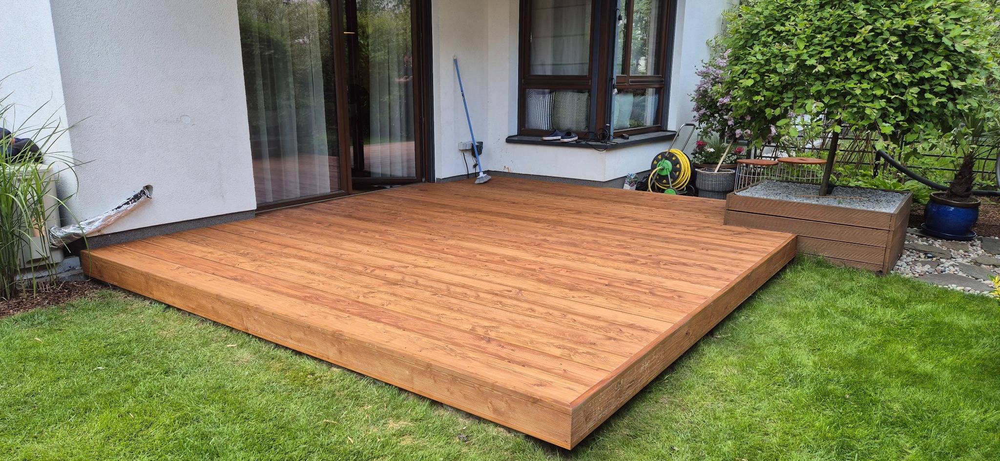 Drewniany taras przy domu, wykonany z desek o ciepłym, brązowym odcieniu. Widok na trawnik, okno i drzewo w donicy. W tle widoczna ściana budynku.