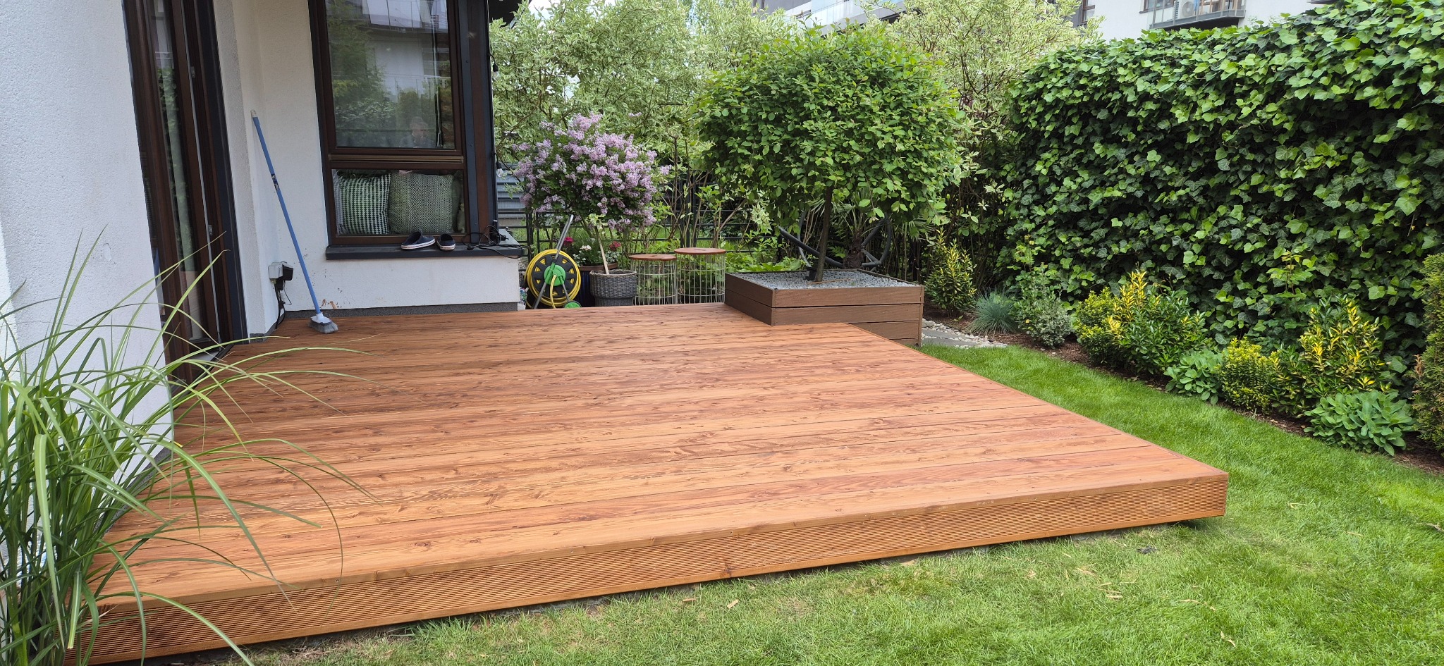 Zrealizowany taras z desek o ciepłym odcieniu drewna, otoczony zielonym trawnikiem i roślinnością. Widoczny fragment domu z oknem i elementami dekoracyjnymi.