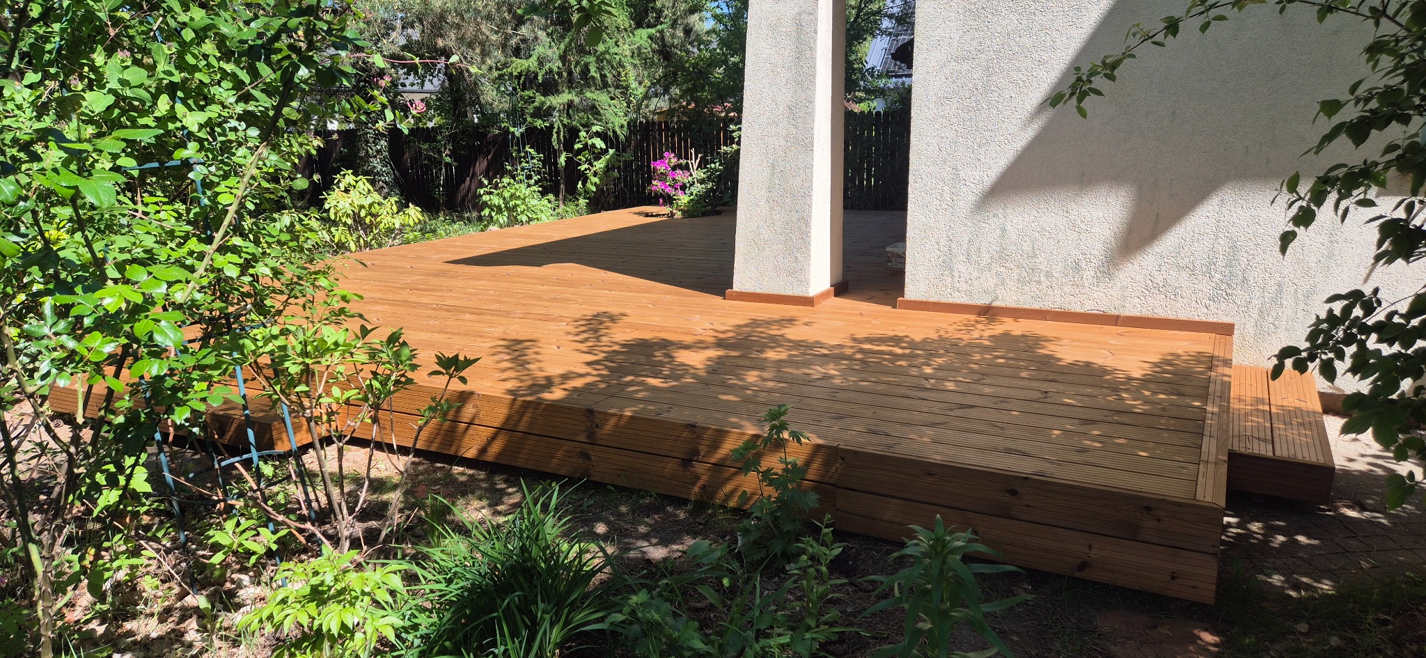 Nowoczesny taras z drewna, z subtelnym oświetleniem, wkomponowany w bujną zieleń ogrodu. Widoczne deski tarasowe i fragment budynku.