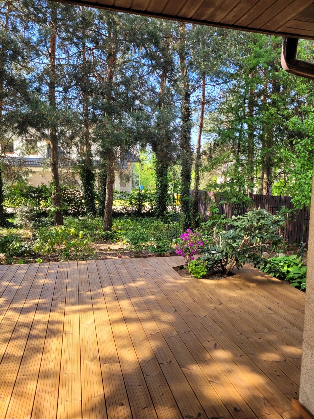 Jasny, drewniany taras z wbudowaną rabatą kwiatową, otoczony zielenią drzew i krzewów. Widok zadaszony, w tle dom.