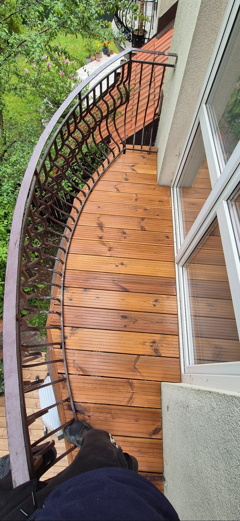 Kątowe ujęcie drewnianego tarasu balkonowego z metalową, dekoracyjną balustradą. Widoczne deski tarasowe i fragment okna. Na podłodze widoczne stopy osoby robiącej zdjęcie.