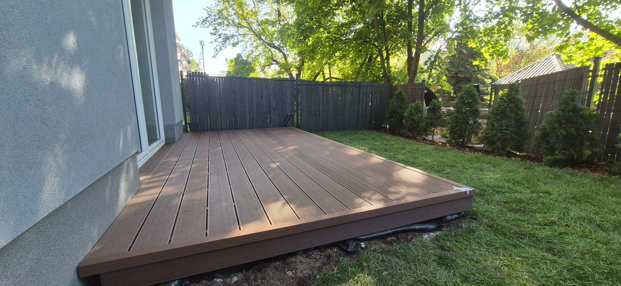 Nowoczesny taras kompozytowy przylegający do domu, z widocznym ogrodzeniem w tle i zielonym trawnikiem, wykonany z desek w odcieniach brązu.