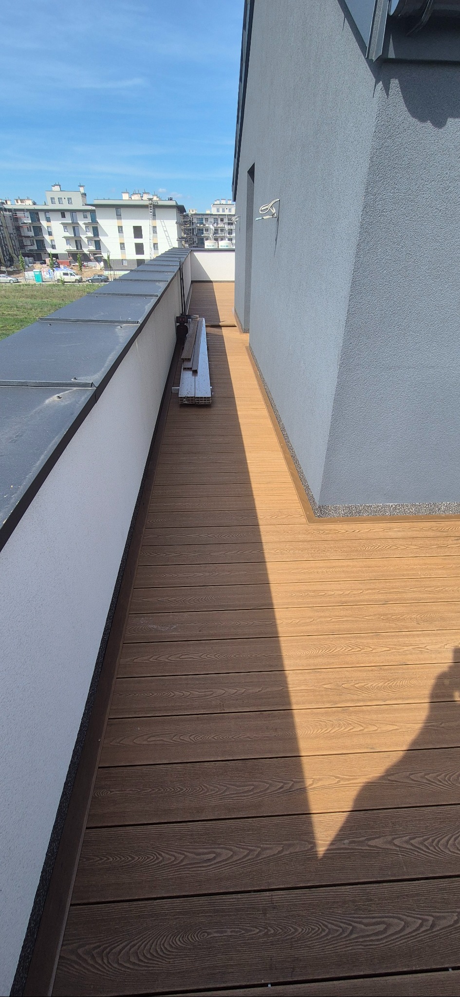 Nowoczesny taras z desek kompozytowych w odcieniach brązu, z widocznymi elementami montażowymi i fragmentem budynku w tle. Widok z góry, słoneczne oświetlenie.