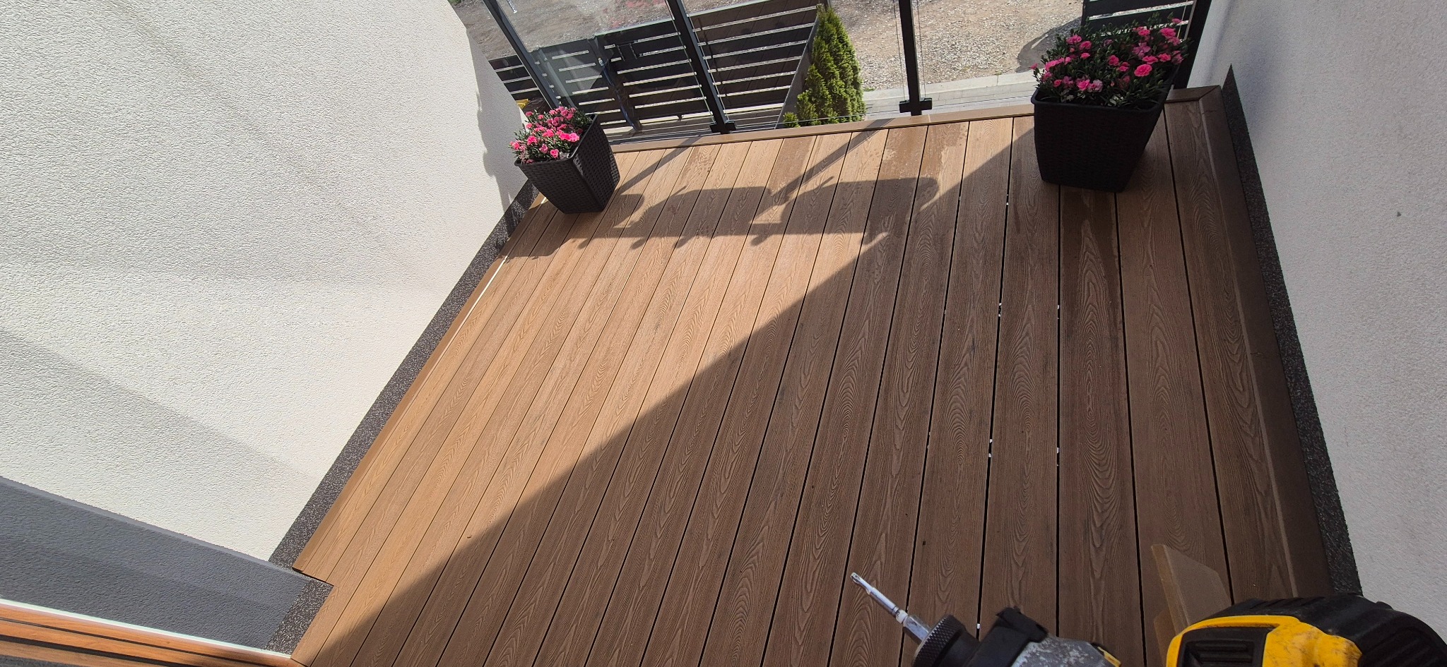 Wykończony taras z desek kompozytowych, z donicami z kwiatami, z narzędziem w rogu. Widok z góry, jasne oświetlenie.