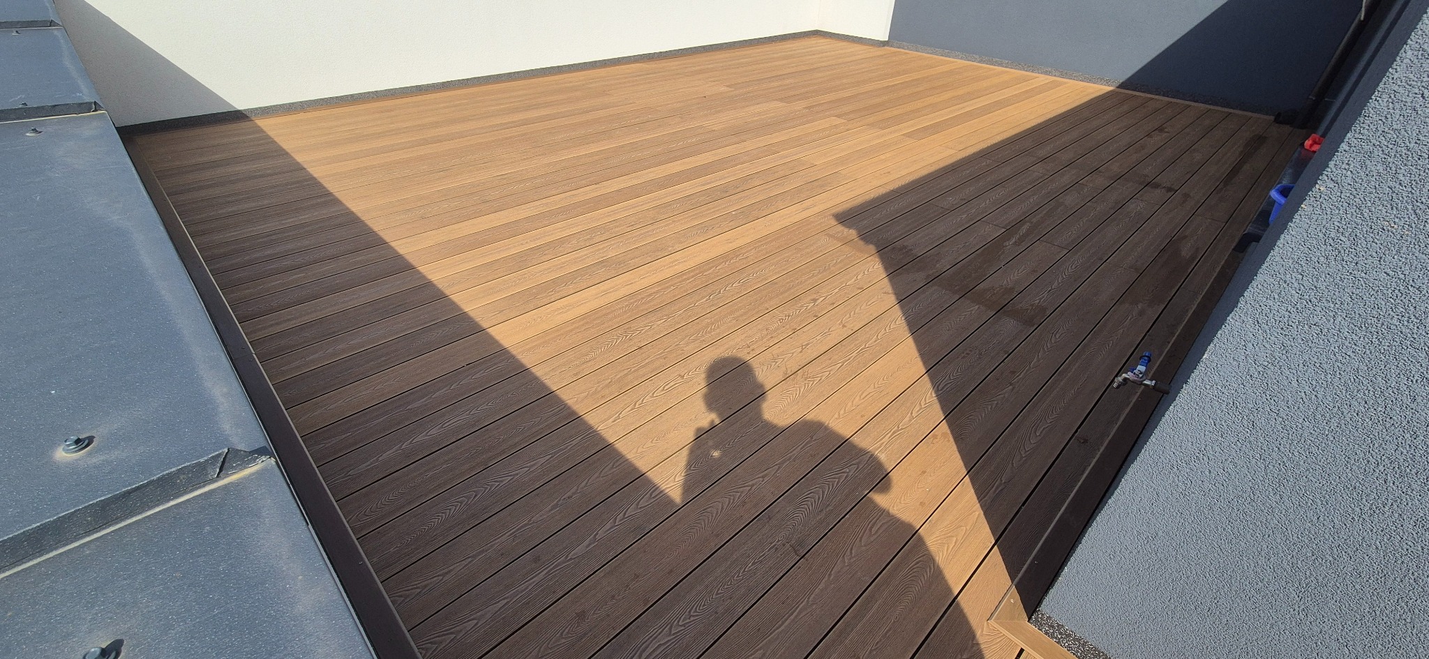 Jasny taras z desek kompozytowych, cień sylwetki osoby, widok z góry. Nowoczesny design, schludne wykończenie, widoczny kran na ścianie.