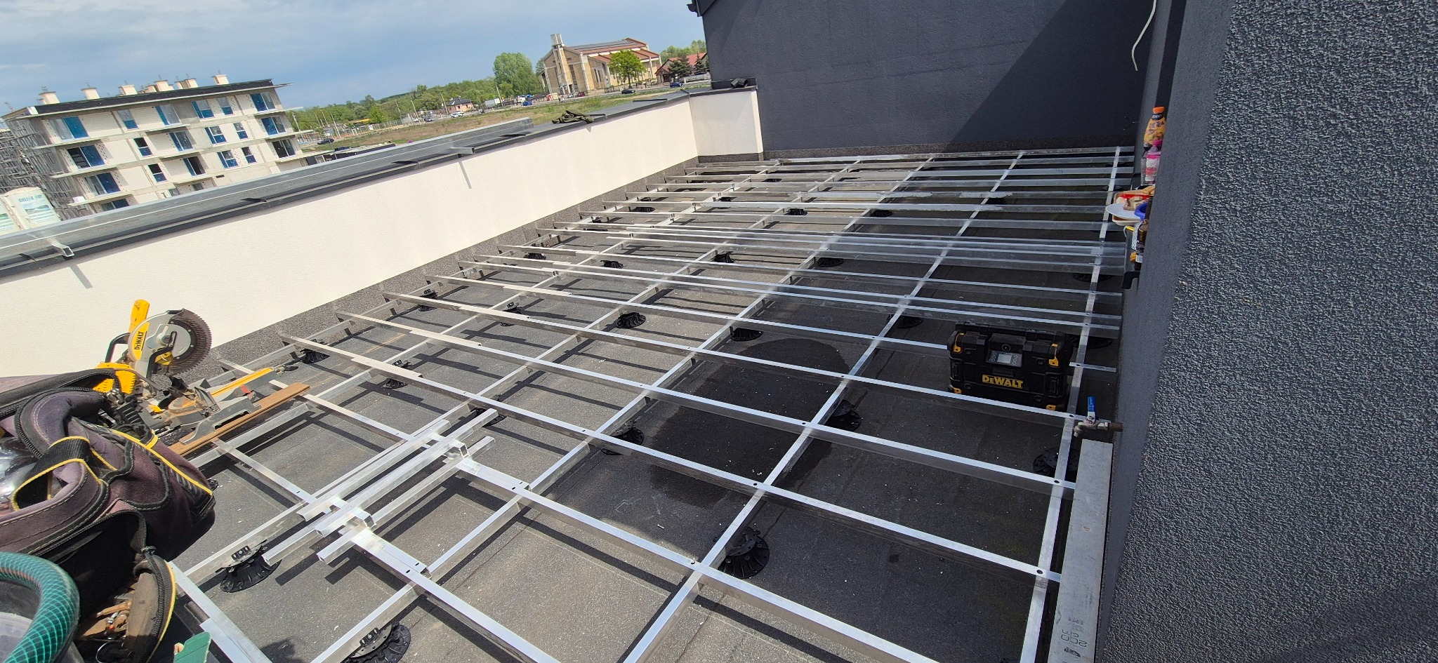 Konstrukcja aluminiowa pod taras kompozytowy na dachu budynku w Makowie Mazowieckim, z narzędziami i skrzynką Dewalt w kadrze. Widok z góry, etap budowy.
