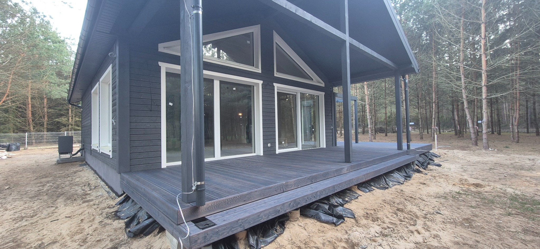 Nowoczesny, ciemny taras drewniany z zadaszeniem, otoczony lasem. Widoczne czarne folie pod konstrukcją. Uwagę przyciągają duże, przesuwne okna.