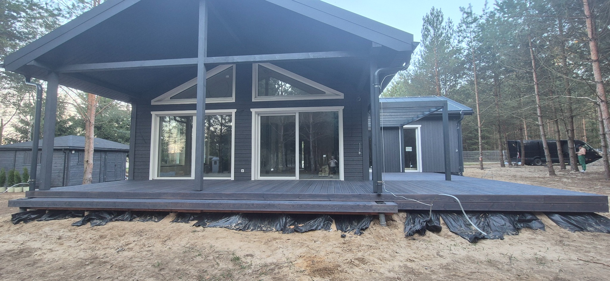 Nowoczesny dom z ciemnego drewna z rozległym tarasem. Widoczne duże okna i otoczenie leśne. Taras w trakcie wykończenia, z widoczną folią ochronną.