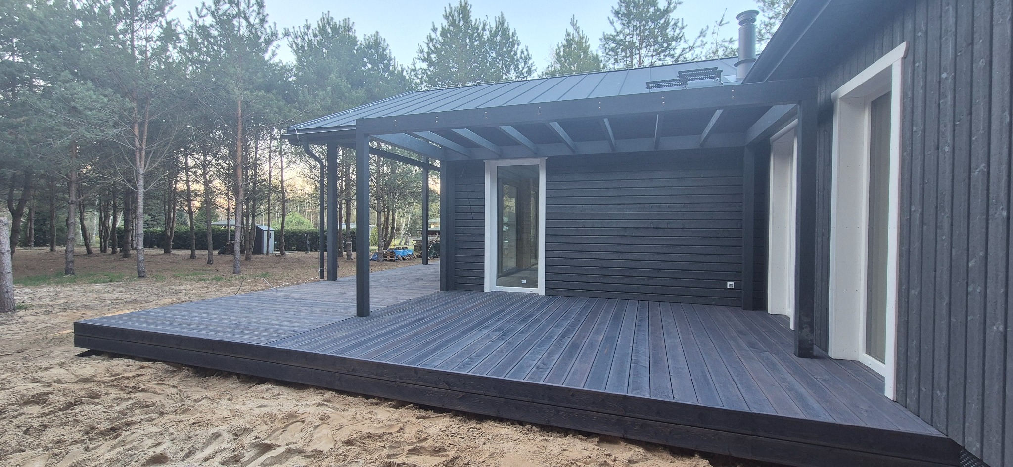 Ciemny, drewniany taras przylegający do nowoczesnego domu z ciemną elewacją i pergolą. W tle las sosnowy i piaszczyste podłoże.
