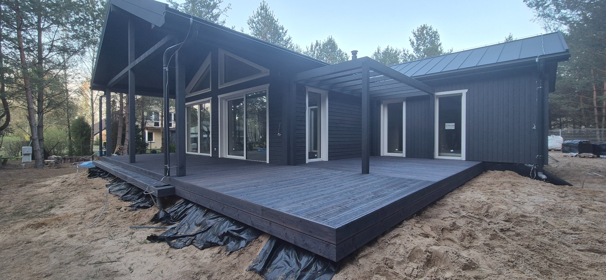 Nowoczesny, ciemny taras z drewna przy domu, otoczony piaskiem i lasem. Widoczna pergola i duże okna przesuwne. Częściowo widoczna folia pod tarasem.