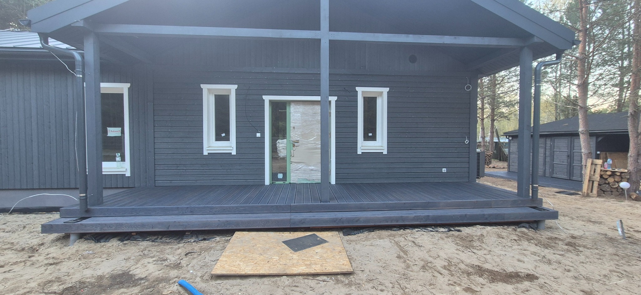 Szara konstrukcja z tarasem w lesie. Widoczne drewniane deski tarasowe i elementy konstrukcyjne. Drzwi zabezpieczone folią. W tle drzewa i budynek gospodarczy.