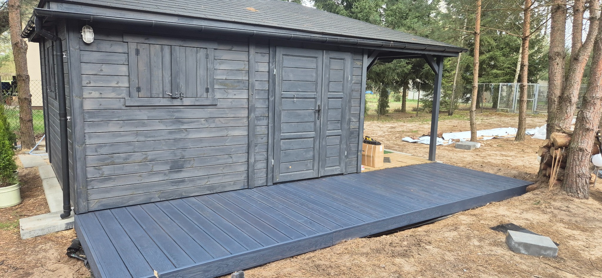 Ciemnoszara, drewniana altana z tarasem, widok z boku. Na tarasie widoczne deski kompozytowe. W tle drzewa i elementy ogrodu.