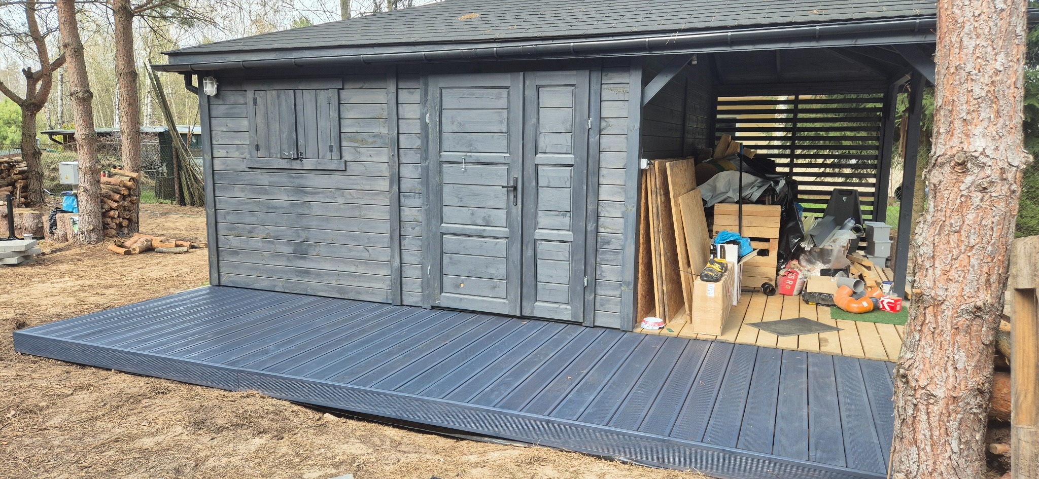 Szara altana z drewnianą podłogą i zadaszeniem, widok z zewnątrz. Na podłodze w altanie widoczne różne przedmioty. Obok altany leżą pnie drzew.