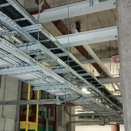 Metalowe korytka kablowe pod stropem budynku w trakcie montażu, widoczne przewody i elementy konstrukcji, w tle kartony z materiałami.