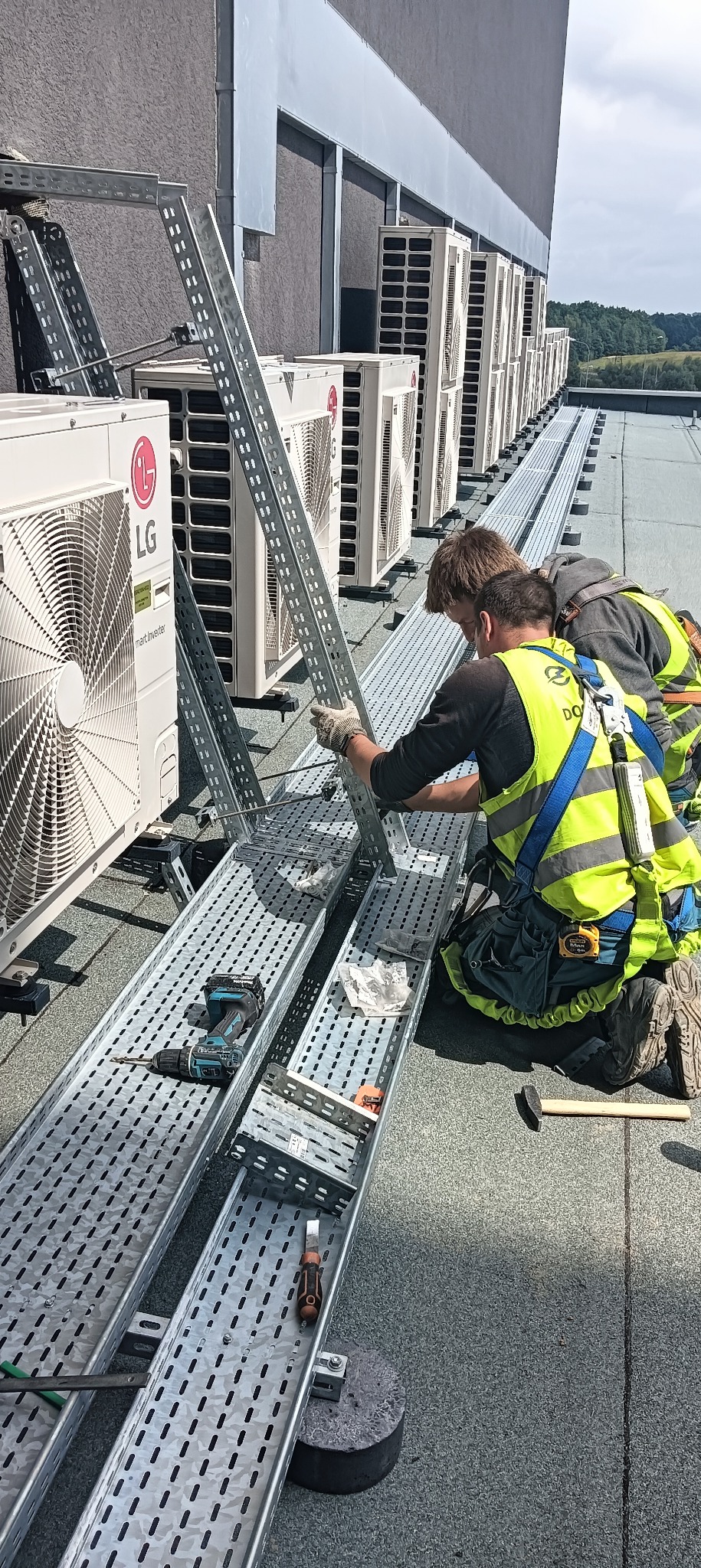 Dwóch monterów w kamizelkach odblaskowych instaluje metalowe korytka kablowe na dachu budynku obok rzędu agregatów klimatyzacyjnych LG, używając wkrętarki akumulatorowej i rękawic roboczych.