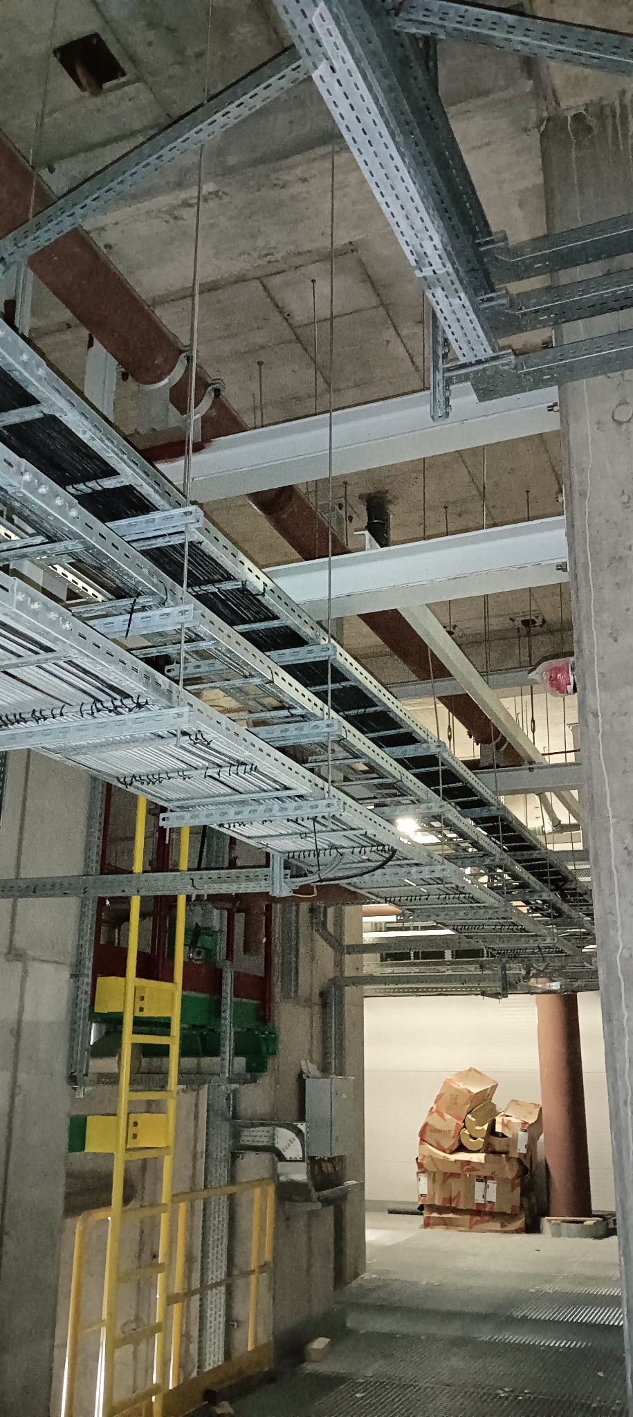 Metalowe korytka kablowe pod stropem budynku w trakcie montażu, widoczne przewody i elementy konstrukcji, w tle kartony z materiałami.