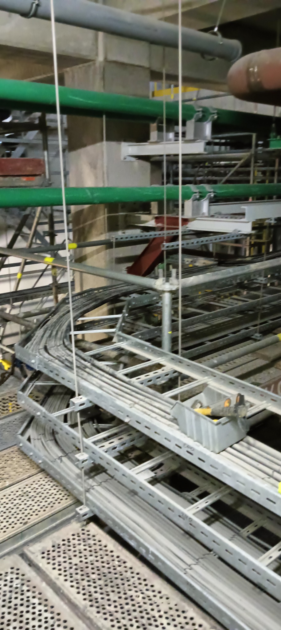 Metalowe korytka kablowe, wypełnione przewodami, zamontowane pod sufitem w hali przemysłowej, widoczne rury i elementy konstrukcyjne budynku.