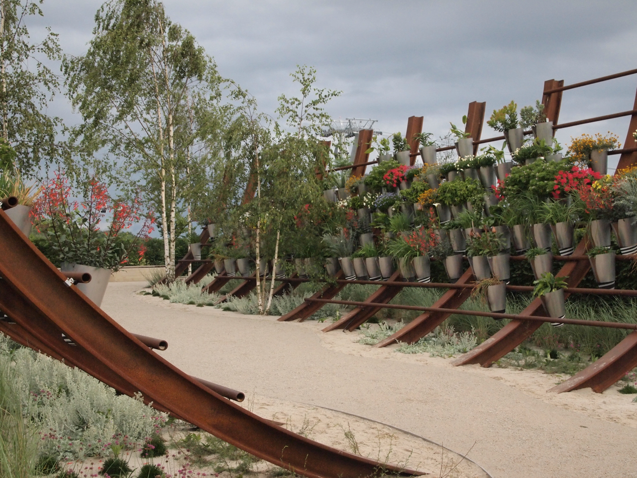 Kaskadowa ściana z roślin w metalowych doniczkach, rustykalna konstrukcja z brązowej stali, żwirowa ścieżka i brzozy w tle, aranżacja ogrodu w stylu naturalnym.