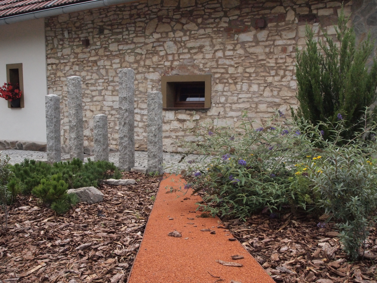 Ogród z elementami dekoracyjnymi: rząd granitowych słupków, ścieżka z czerwonej nawierzchni, rabaty z korą, krzewami i kwiatami na tle kamiennej ściany budynku.