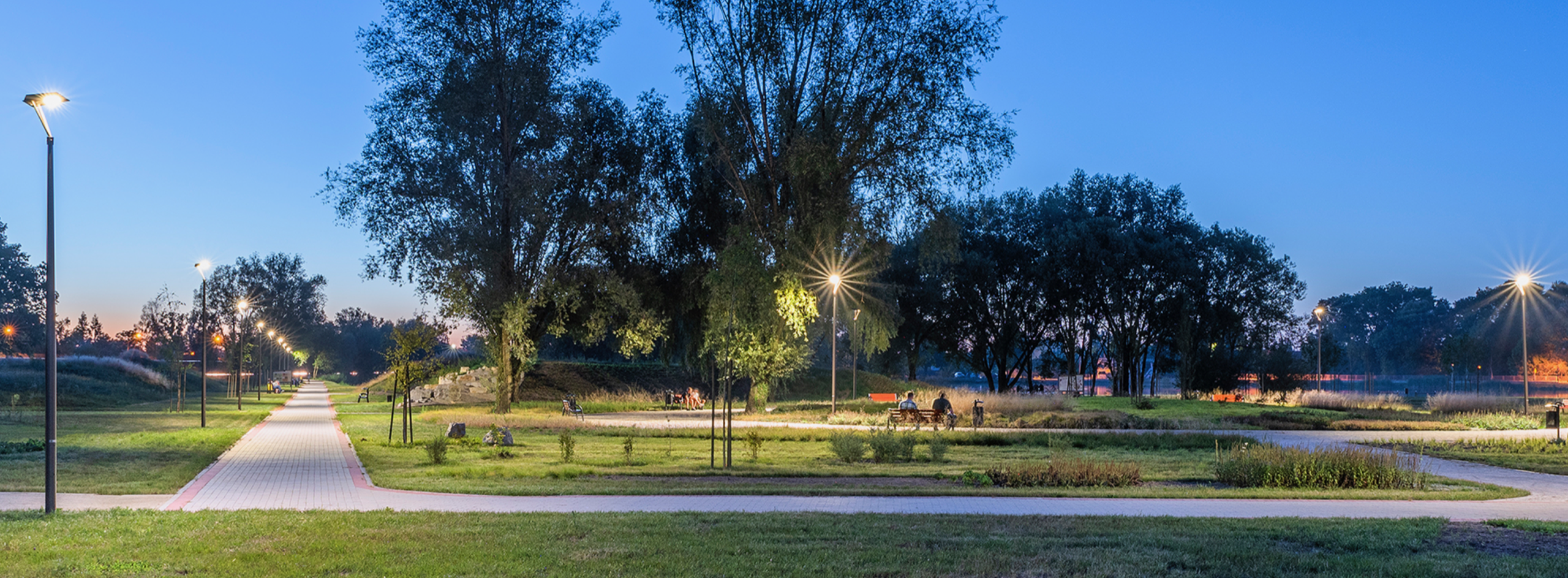 Oświetlony park miejski o zmierzchu, z alejkami wyłożonymi kostką, ławkami i drzewami, widok na oświetlone latarniami ścieżki i sylwetki ludzi siedzących na ławkach w oddali.