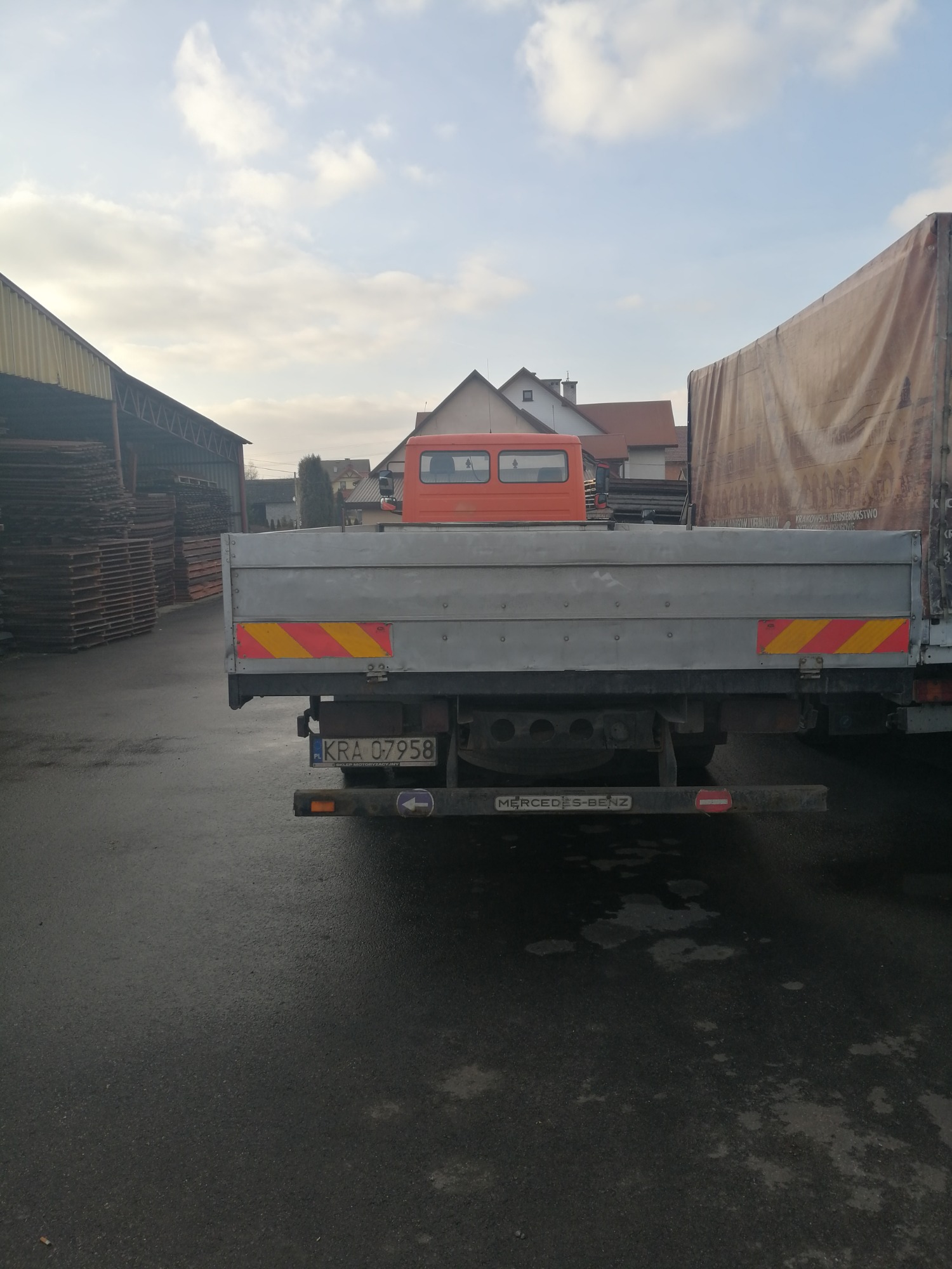 Tył szarego samochodu ciężarowego Mercedes-Benz z widoczną tablicą rejestracyjną KRA 07958 na mokrym asfalcie, z budynkiem magazynowym i stertą drewna w tle.