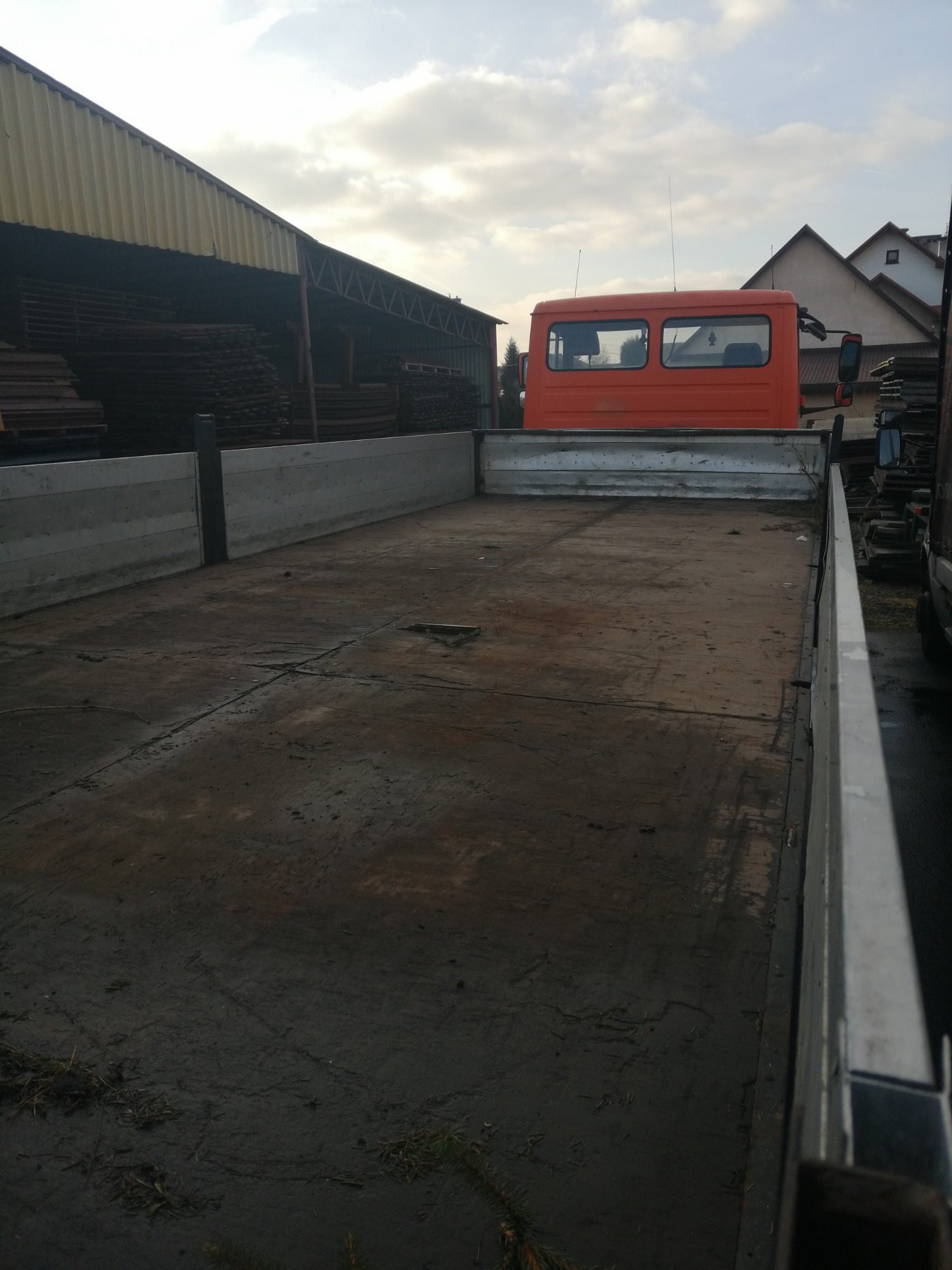 Pusta skrzynia ładunkowa pomarańczowego samochodu ciężarowego z widocznymi śladami użytkowania, na tle zabudowań gospodarczych i fragmentu dachu domu.