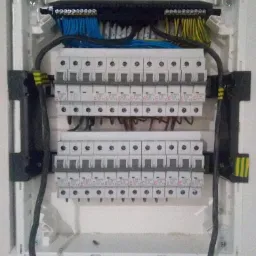 Rozdzielnica elektryczna natynkowa z dwoma rzędami wyłączników, okablowaniem w kolorach niebieskim, żółto-zielonym i czarnym, widoczne oznaczenia na wyłącznikach.