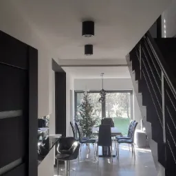 Wnętrze domu z widoczną kuchnią z barkiem, jadalnią z czarnymi krzesłami i schodami w ciemnej tonacji, oświetlone punktowymi lampami sufitowymi i naturalnym światłem wpadającym przez okno,...