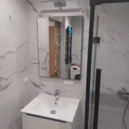 Nowoczesna łazienka w Mysłowicach z marmurowymi płytkami, białą szafką pod umywalką i czarną kabiną prysznicową. W lustrze odbija się fragment ściany w trakcie prac oraz pralka.