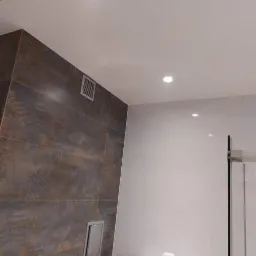 Nowoczesna łazienka z brązowymi płytkami imitującymi drewno, białymi ścianami, punktowym oświetleniem sufitowym i szklaną kabiną prysznicową. Na blacie umywalki widoczny kubek po kawie i drobne...