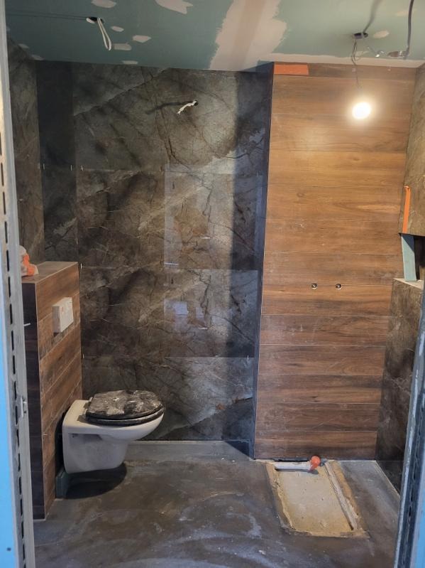 Łazienka w trakcie remontu: ściany wyłożone płytkami imitującymi marmur i drewno, widoczny fragment toalety z zabrudzoną deską, surowy sufit i podłoga w trakcie prac wykończeniowych.