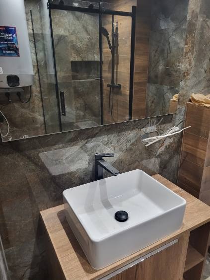 Nowoczesna łazienka z kwadratową umywalką nablatową, czarną armaturą i kabiną prysznicową typu walk-in z ciemnymi płytkami imitującymi kamień.