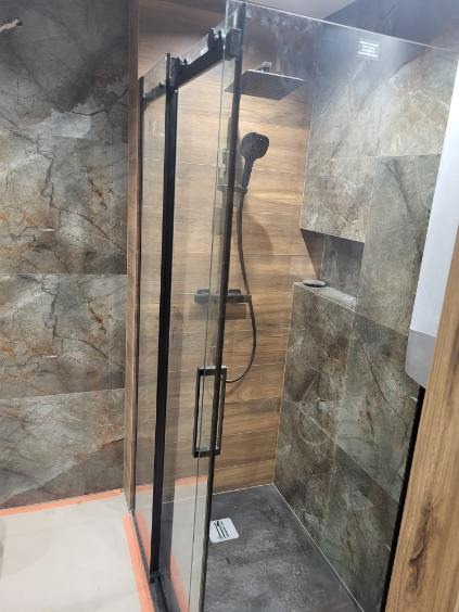 Nowoczesna kabina prysznicowa z czarnymi profilami i deszczownicą, wykończona płytkami imitującymi kamień w odcieniach brązu i szarości, z widocznym odpływem liniowym.