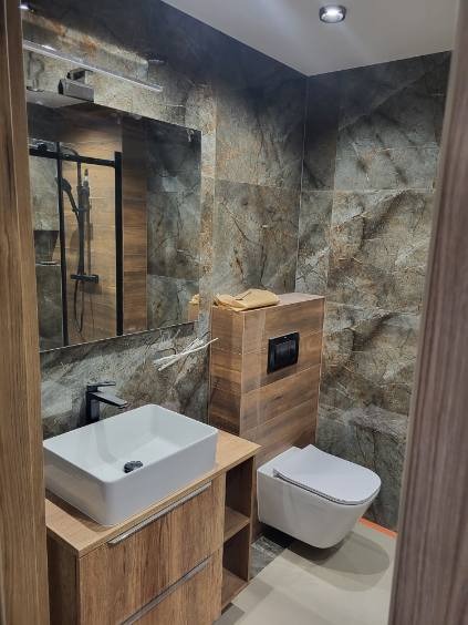 Nowoczesna łazienka z marmurowymi płytkami, wiszącą toaletą i umywalką nablatową na drewnianej szafce, widoczna kabina prysznicowa z czarną ramą w lustrze.