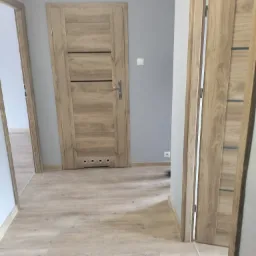 Korytarz z dwiema zamontowanymi, drewnianymi drzwiami wewnętrznymi z poziomymi, ciemniejszymi paskami, jasną podłogą i szarą ścianą z włącznikiem.