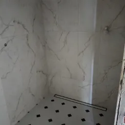 Wykończony brodzik prysznicowy z odpływem liniowym, wyłożony płytkami imitującymi marmur na ścianach i szarymi płytkami z czarnymi wstawkami na podłodze, widoczny fragment nieotynkowanej ściany.