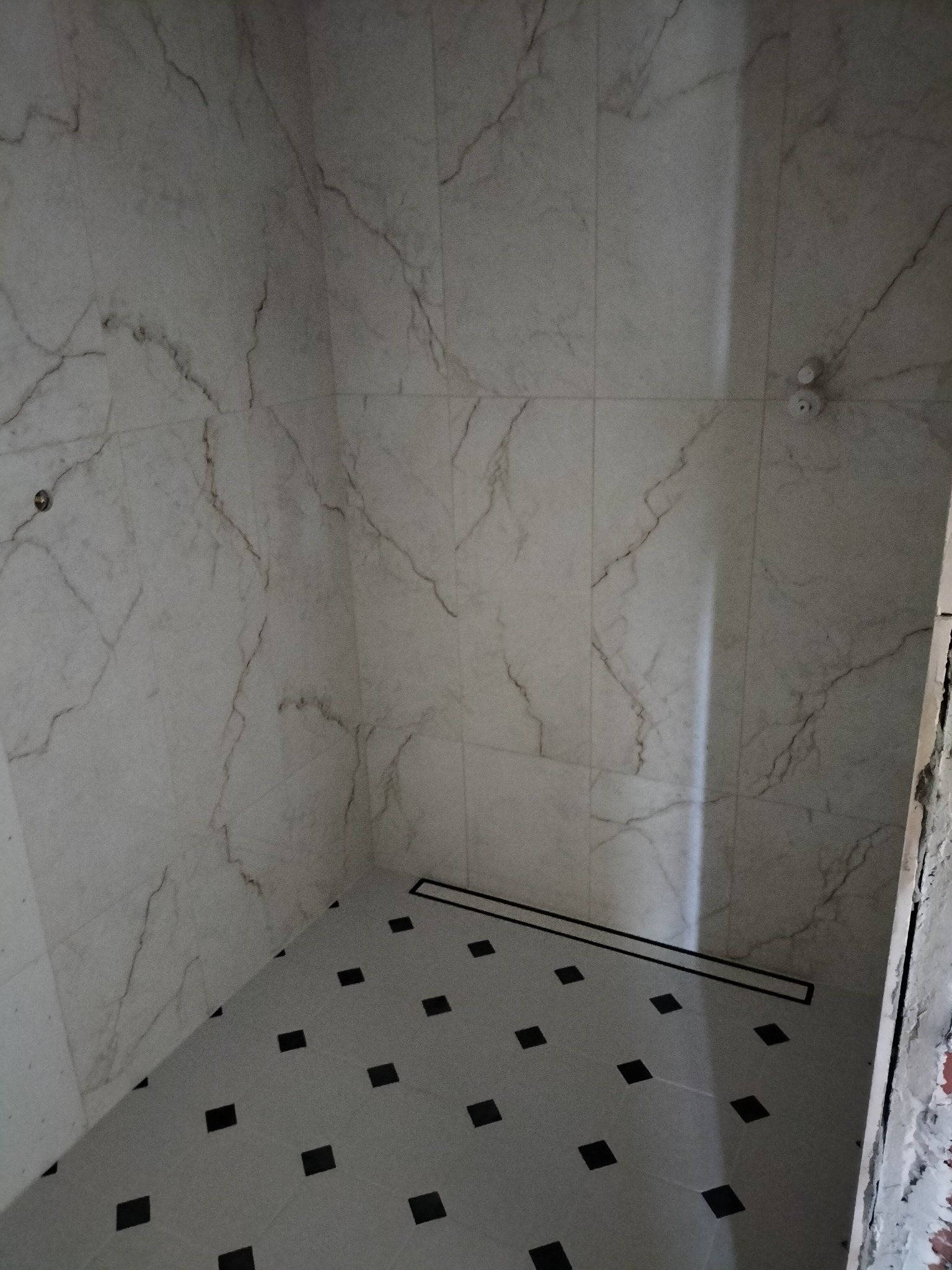 Wykończony brodzik prysznicowy z odpływem liniowym, wyłożony płytkami imitującymi marmur na ścianach i szarymi płytkami z czarnymi wstawkami na podłodze, widoczny fragment nieotynkowanej ściany.