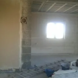 Wnętrze pomieszczenia w trakcie prac tynkarskich: ściana pokryta świeżym tynkiem, widoczny otwór wentylacyjny w rogu, drabina malarska i wiadro na podłodze, sufit z odsłoniętą konstrukcją.