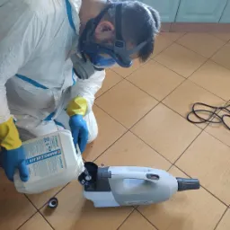 Pracownik w kombinezonie ochronnym i masce wlewa płyn dezynfekujący Nanoclean Air do zamgławiacza na brązowej podłodze z płytek.