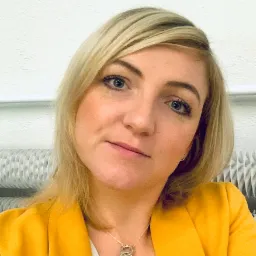 Portret uśmiechniętej blondynki w żółtej marynarce, z widocznym srebrnym naszyjnikiem z zawieszką w kształcie misia, na tle białej ściany z grzejnikiem.