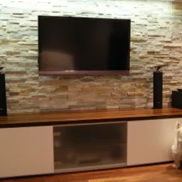 Telewizor na ścianie wyłożonej dekoracyjnym kamieniem, pod nim szafka RTV z drewnianym blatem i białą obudową, obok siedzi biały pies rasy West Highland White Terrier.